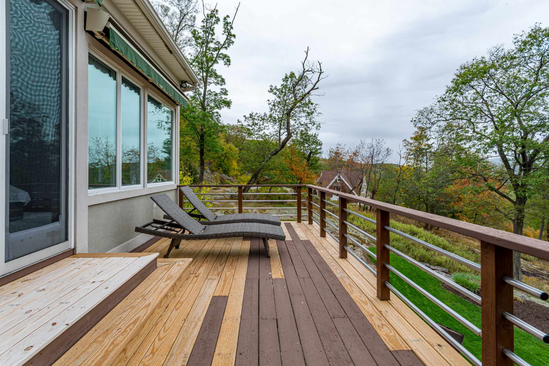 Outdoor wooden deck with two grey lounge chairs, featuring a modern railing with horizontal metal bars and a scenic view of trees and distant hills.