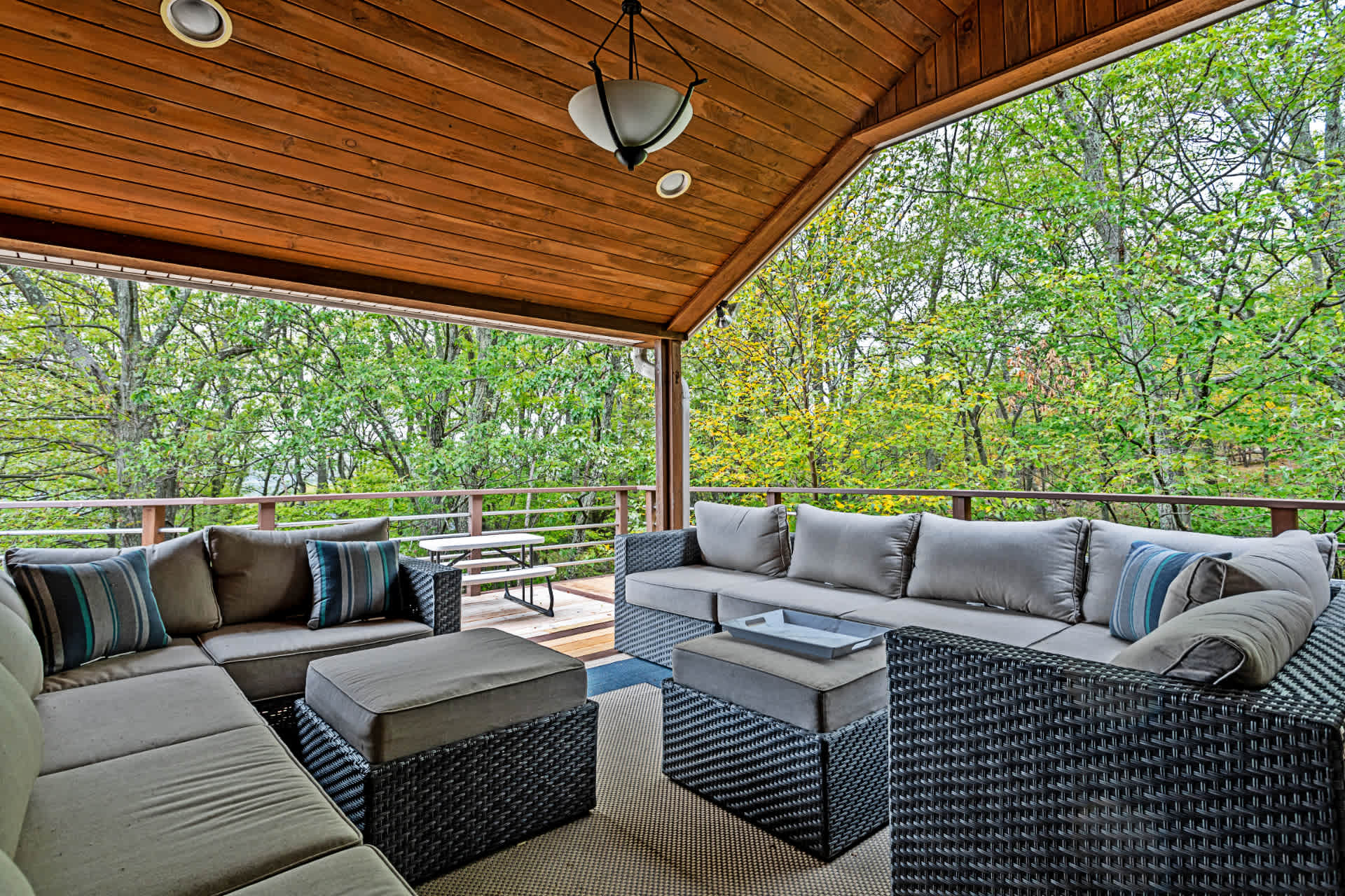 Covered outdoor deck featuring a large L-shaped wicker sectional sofa with gray cushions and blue striped pillows, set on a patterned rug. The wooden ceiling and railing are visible, with lush green trees providing a natural backdrop.