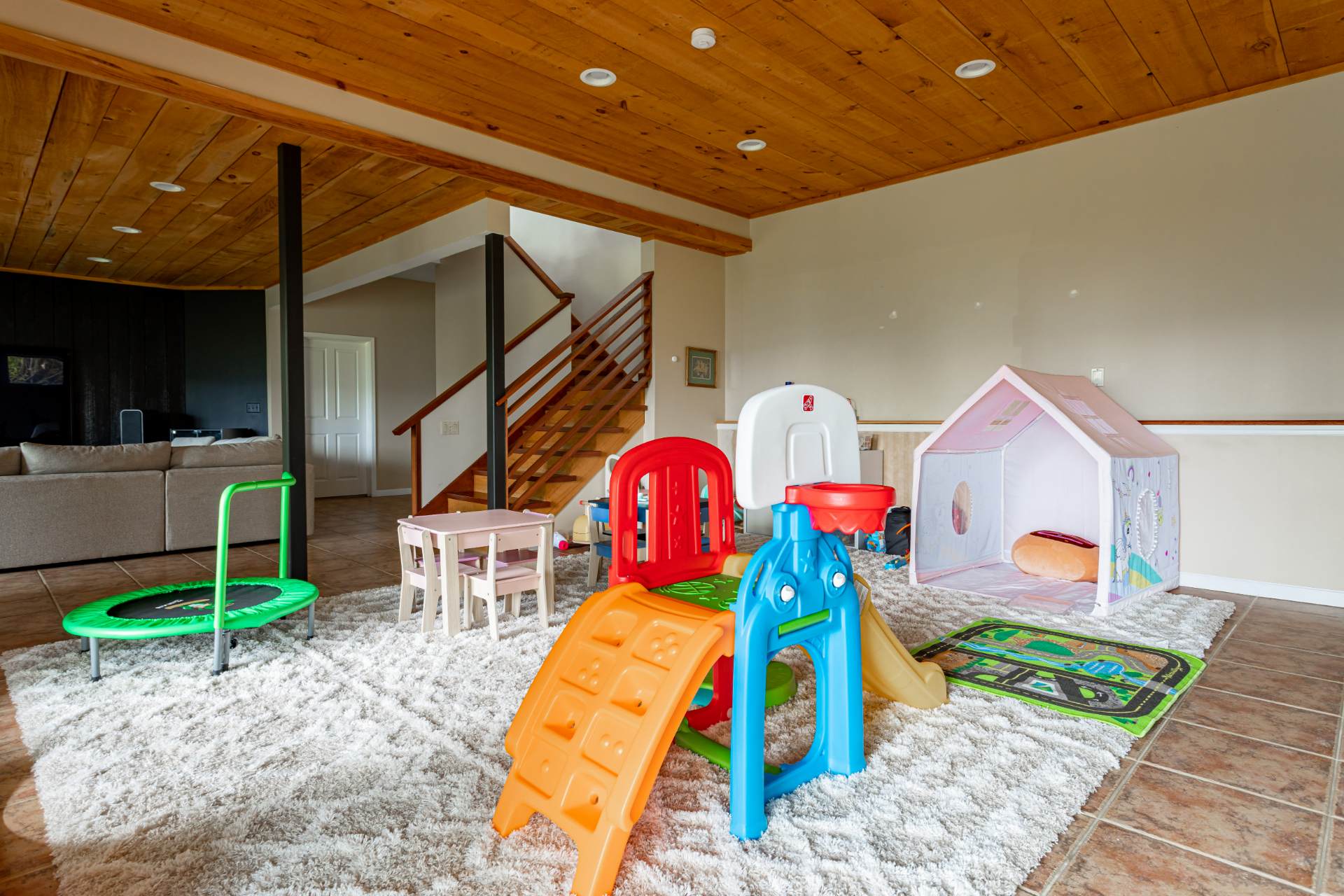 Children's play area with a large white shag rug, various colorful toys including a slide, trampoline, and play tent. The room has a warm wooden ceiling, tiled floors, and a staircase leading to another level.