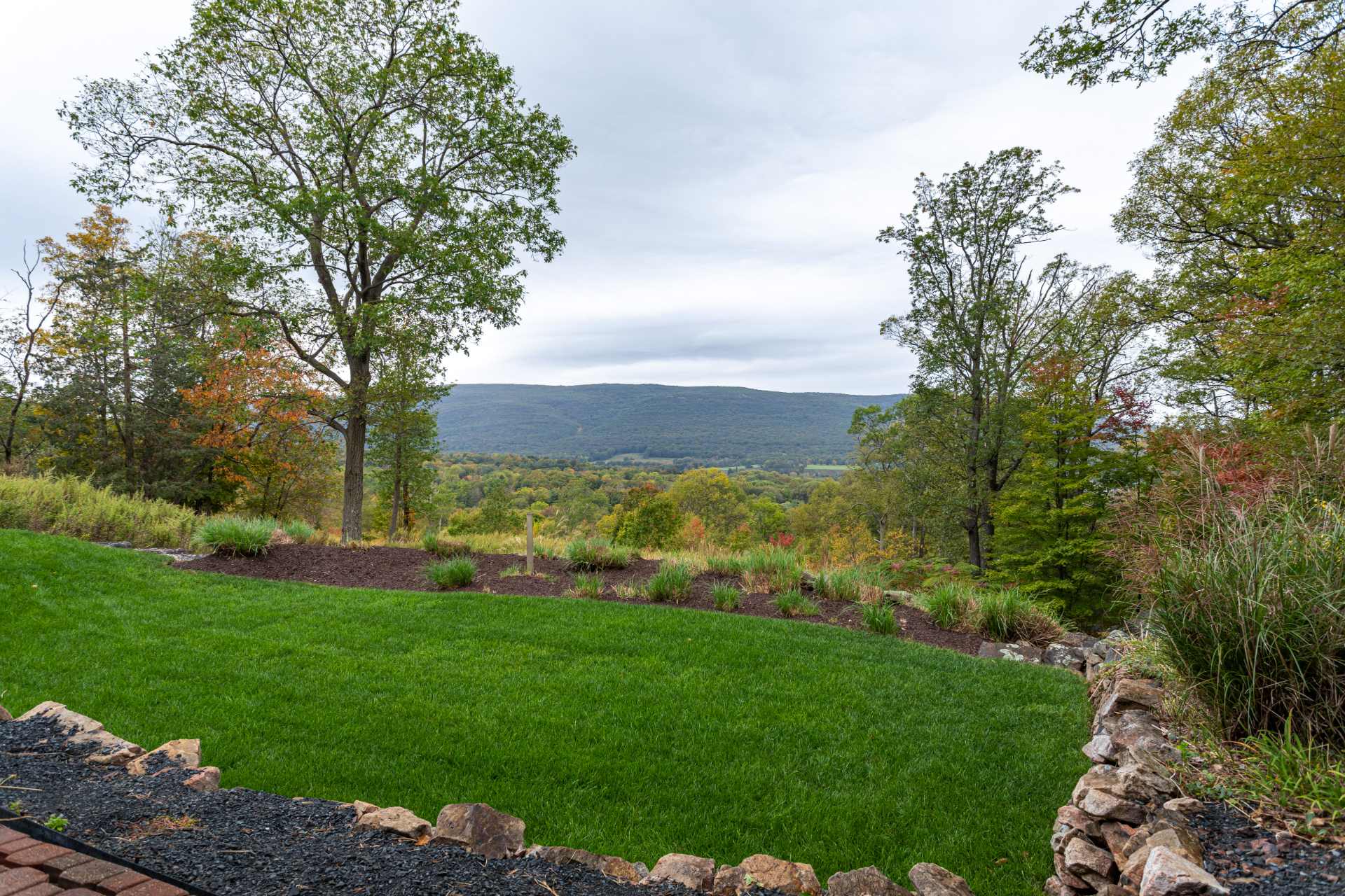 Lush green lawn and landscaped garden area with a low stone wall, offering a breathtaking panoramic view of rolling hills and a distant mountain range under a cloudy sky.