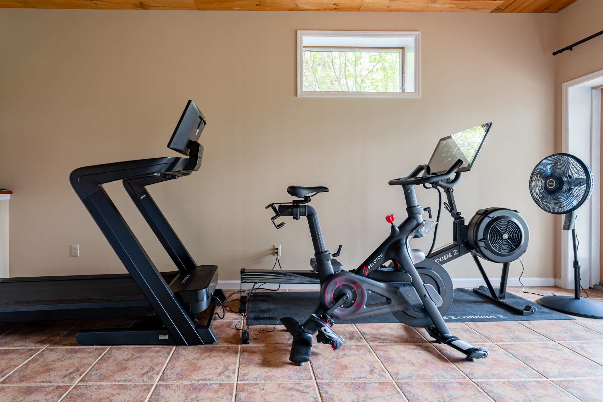 Dedicated fitness corner within a recreation room, showcasing a modern treadmill, a Peloton stationary bike, and a rowing machine on a protective mat.