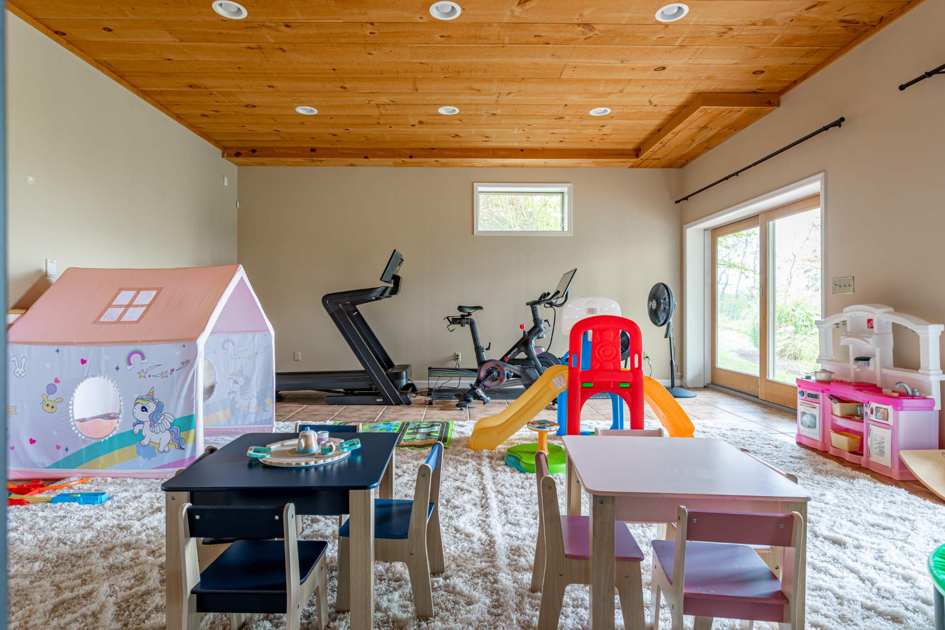 Versatile recreation room featuring a children's play area with small tables and a tent, alongside exercise equipment including a treadmill and stationary bike, all under a wood plank ceiling.