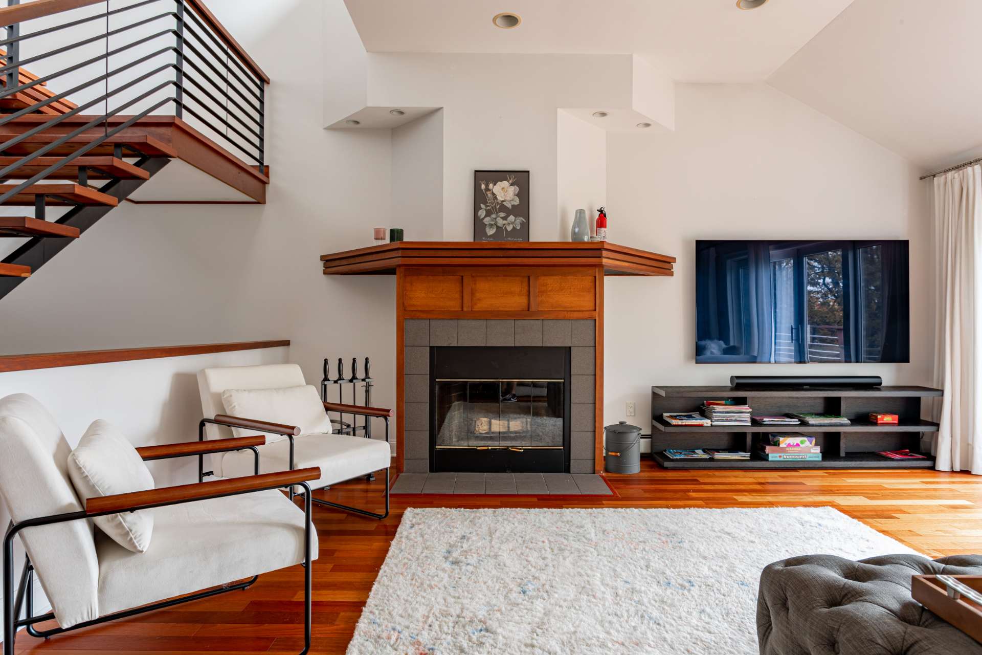 Cozy living room with a prominent fireplace featuring a wooden mantel and gray tile surround, flanked by two white armchairs, set on a plush white rug over hardwood floors.