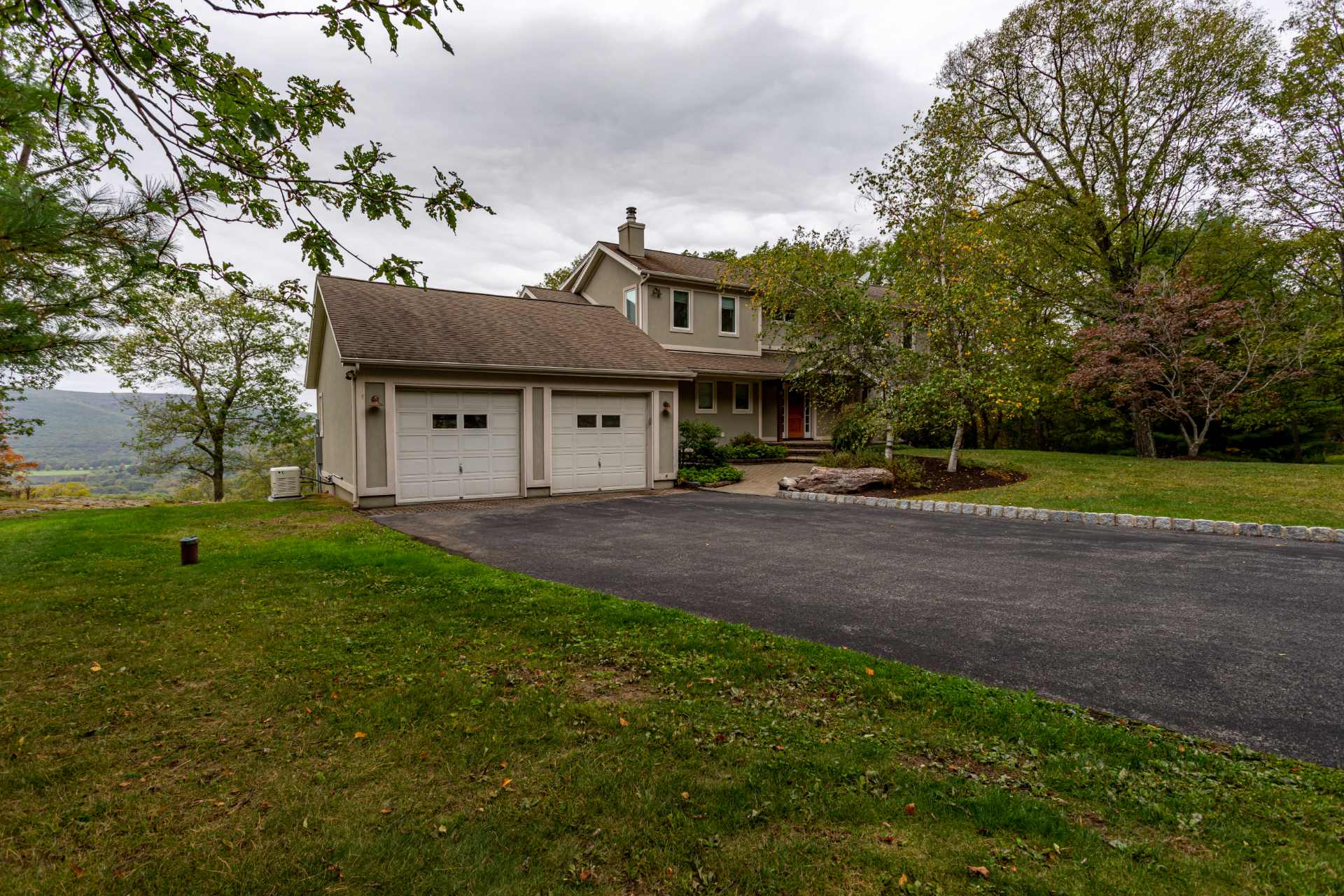 The front exterior of a two-story house with a light beige facade and a two-car attached garage, featuring a paved driveway and a distant mountain view under a cloudy sky.
