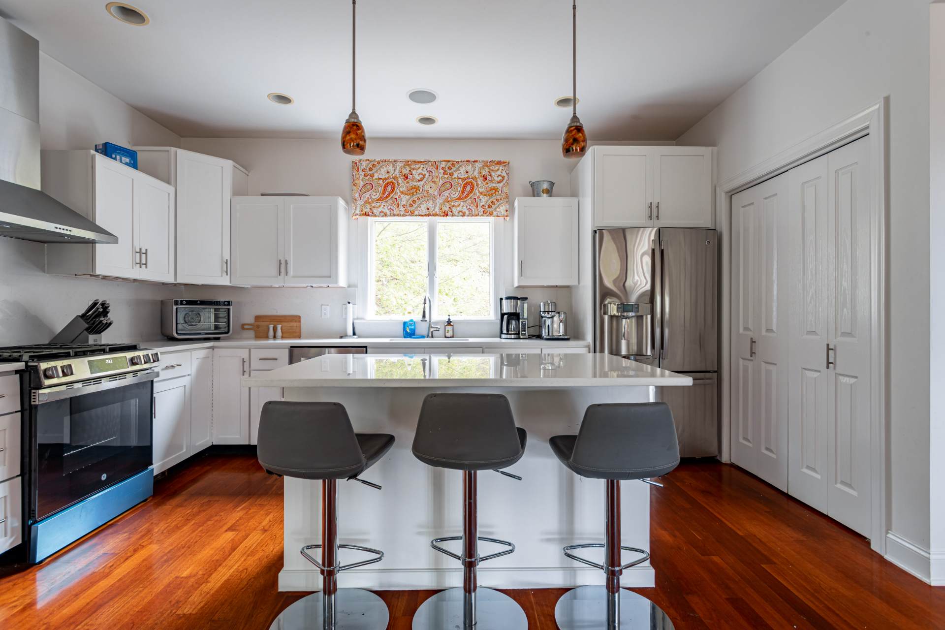 Well-lit kitchen with white shaker cabinets, a large white quartz island with three bar stools, stainless steel appliances, and a window above the sink with a patterned valance.
