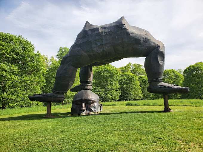 A large, bronze-toned sculpture depicting an inverted human-like figure with its head on the ground and legs arched upwards, set on a lush green lawn surrounded by trees under a bright sky.
