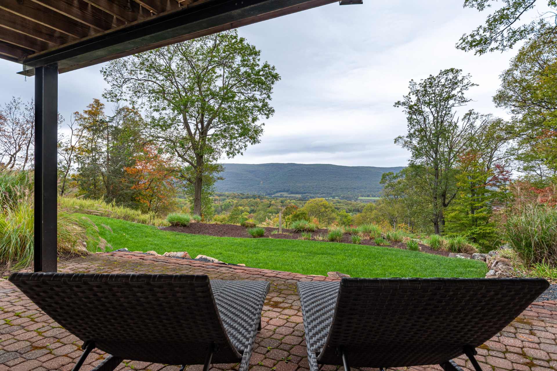 Ground-level brick patio with two lounge chairs, offering stunning panoramic views of a lush green valley and distant mountains. The patio is surrounded by manicured grass and natural rock landscaping.