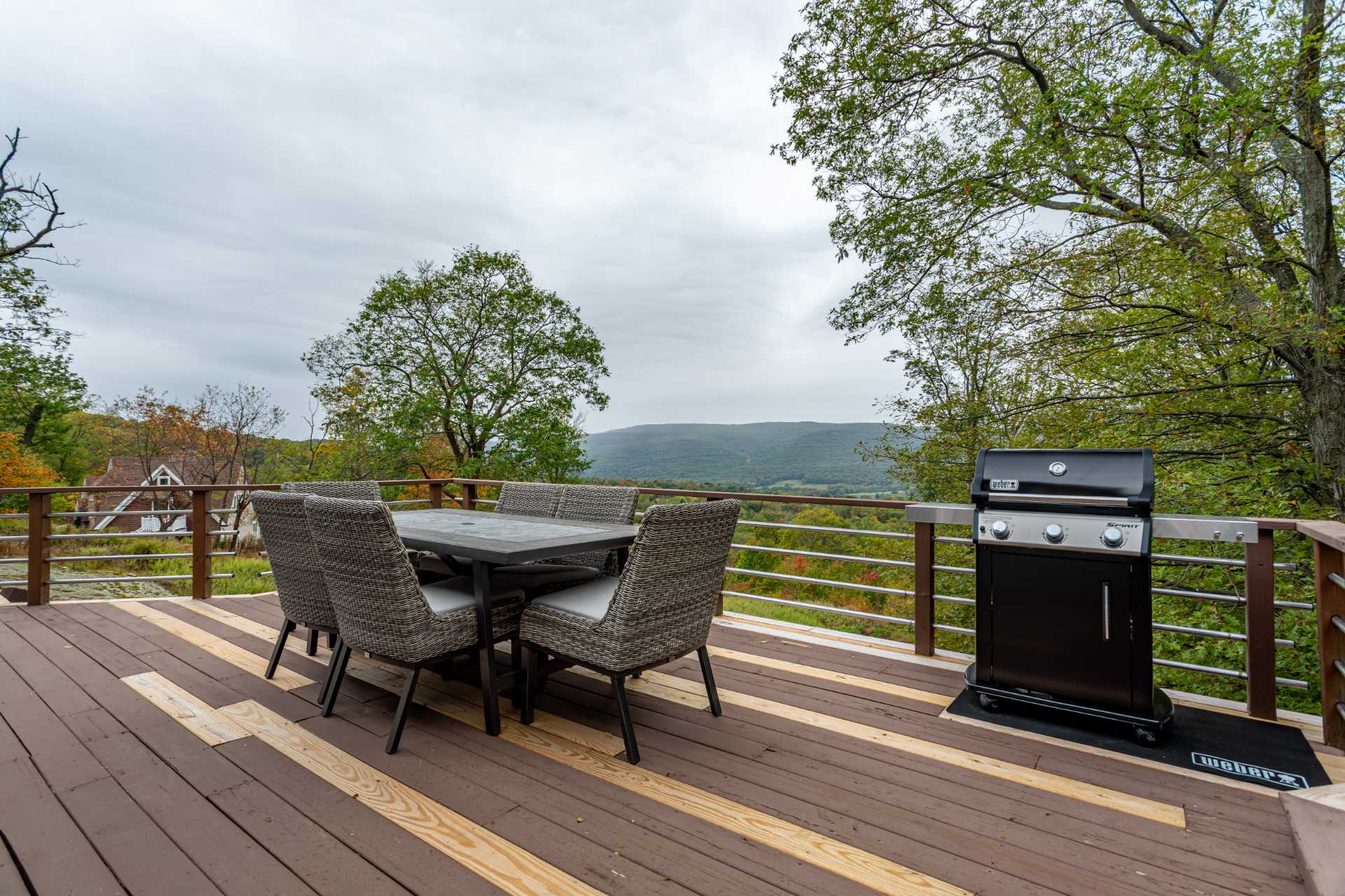 Open wooden deck with an outdoor dining table and chairs, and a BBQ grill, offering expansive views of distant mountains and a valley filled with trees. The deck features a mix of dark and light wood planks and a metal railing.