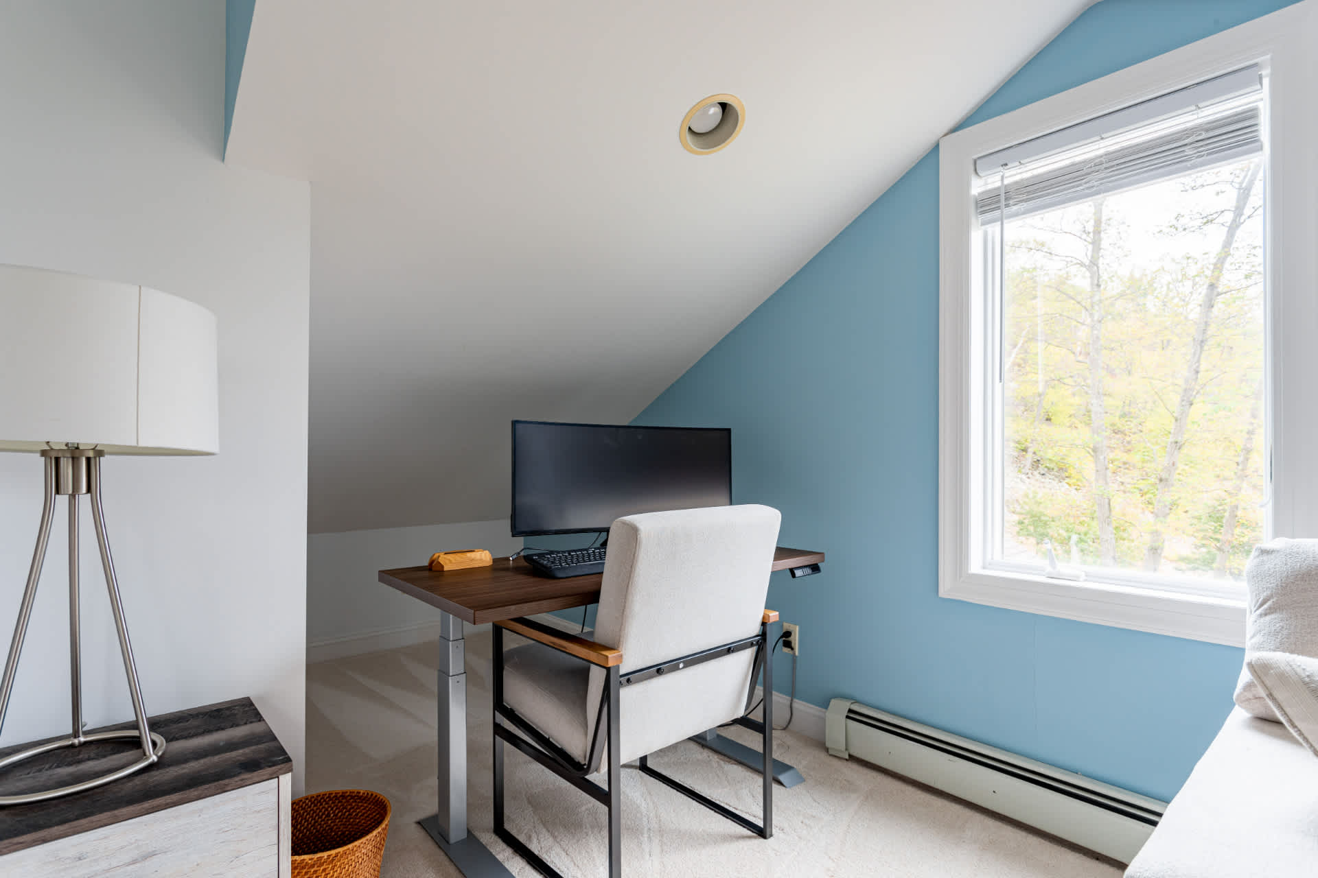 A dedicated workspace within a bedroom, featuring a modern standing desk with a computer monitor and a comfortable office chair, set against a light blue accent wall. A window offers natural light and views of trees.