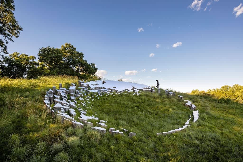 An outdoor art installation featuring a large, circular arrangement of rough-hewn stone blocks on a grassy hillside, partially covered in snow, under a bright blue sky with a person standing in the distance.