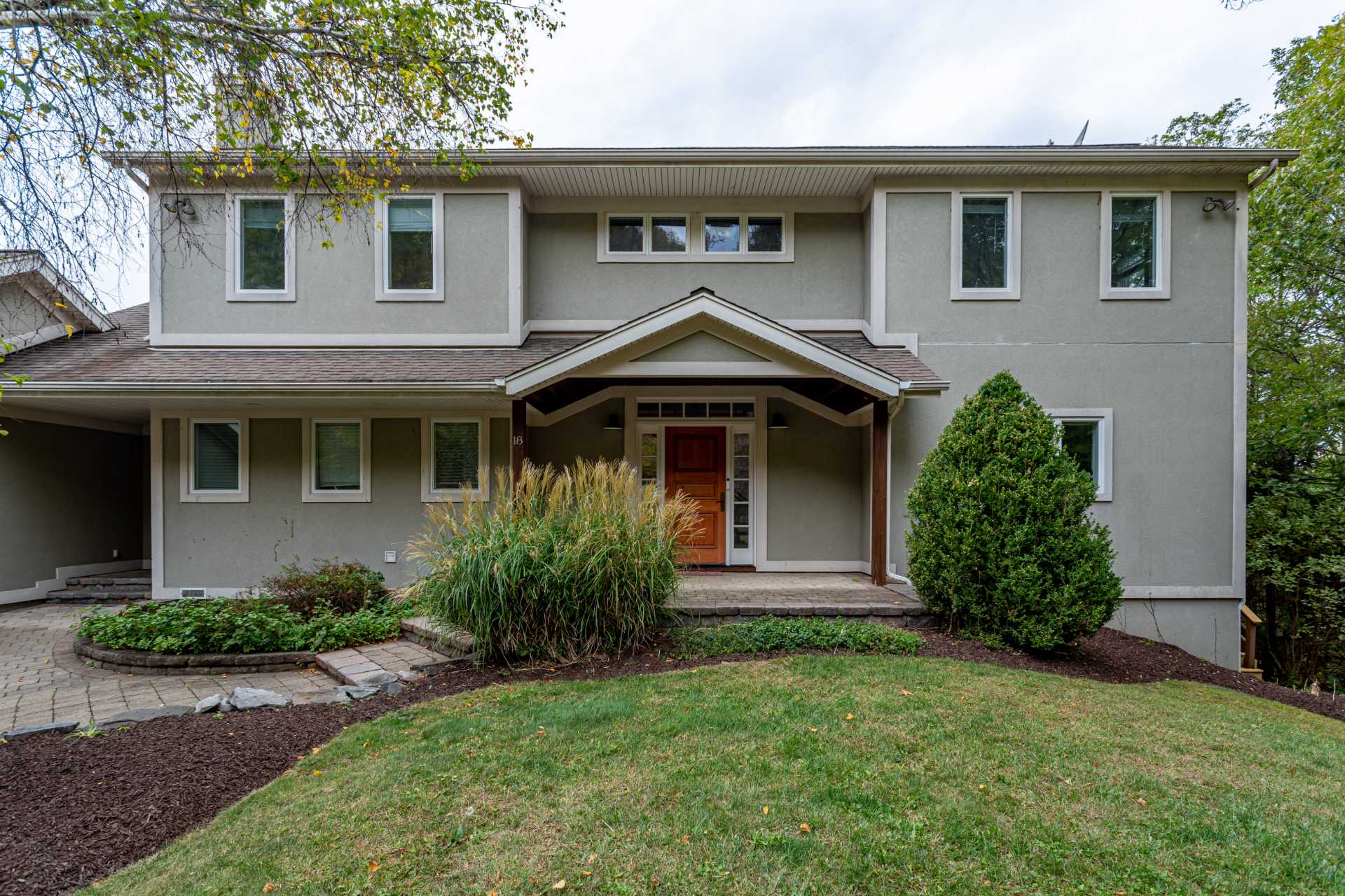 The front exterior of a modern grey house with a covered entryway featuring a warm wooden door and columns, surrounded by green landscaping and mature trees. The house has multiple windows with white trim and a paved walkway.
