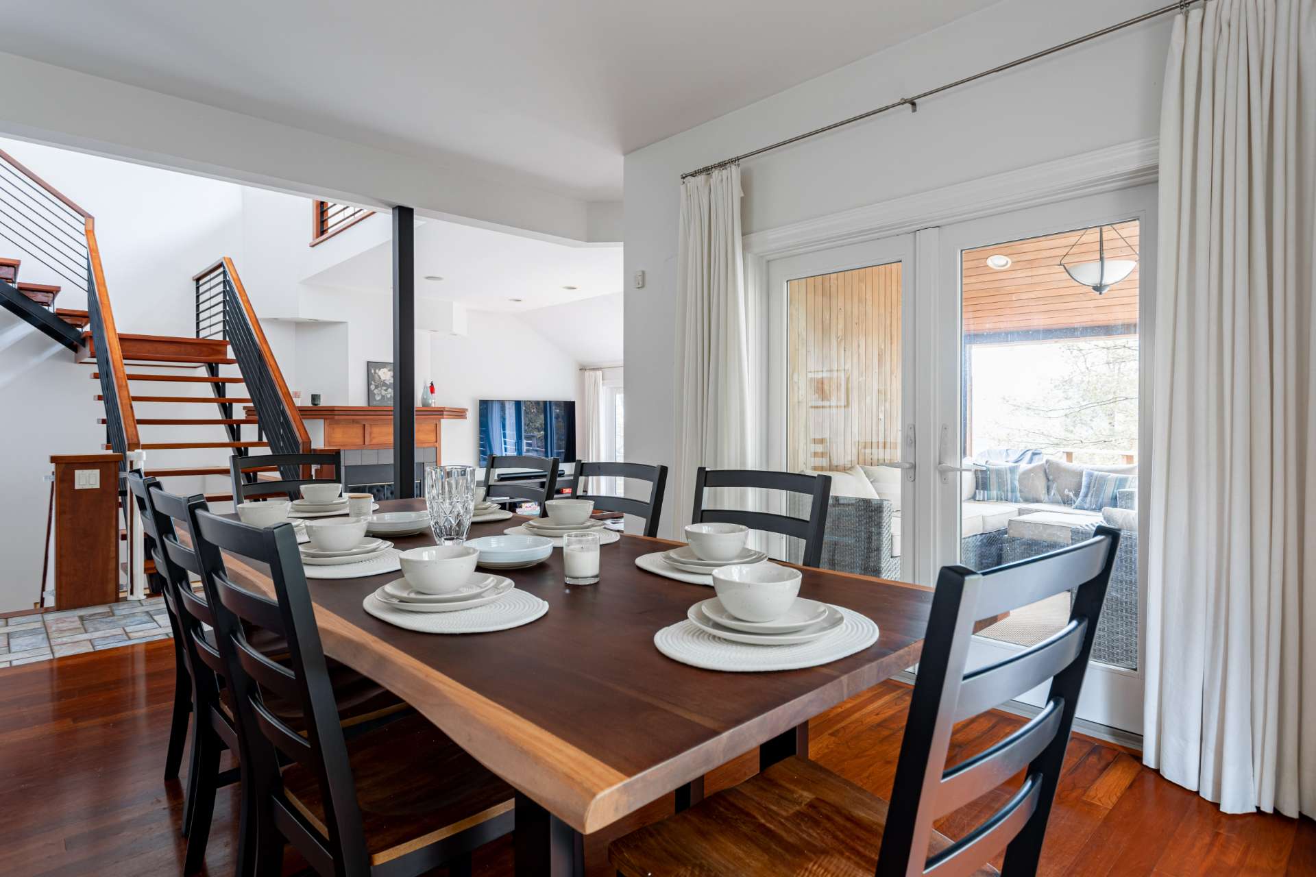 Elegant dining area featuring a dark wooden table set for eight with white place settings, adjacent to sliding glass doors and offering an open view to the living room and staircase.