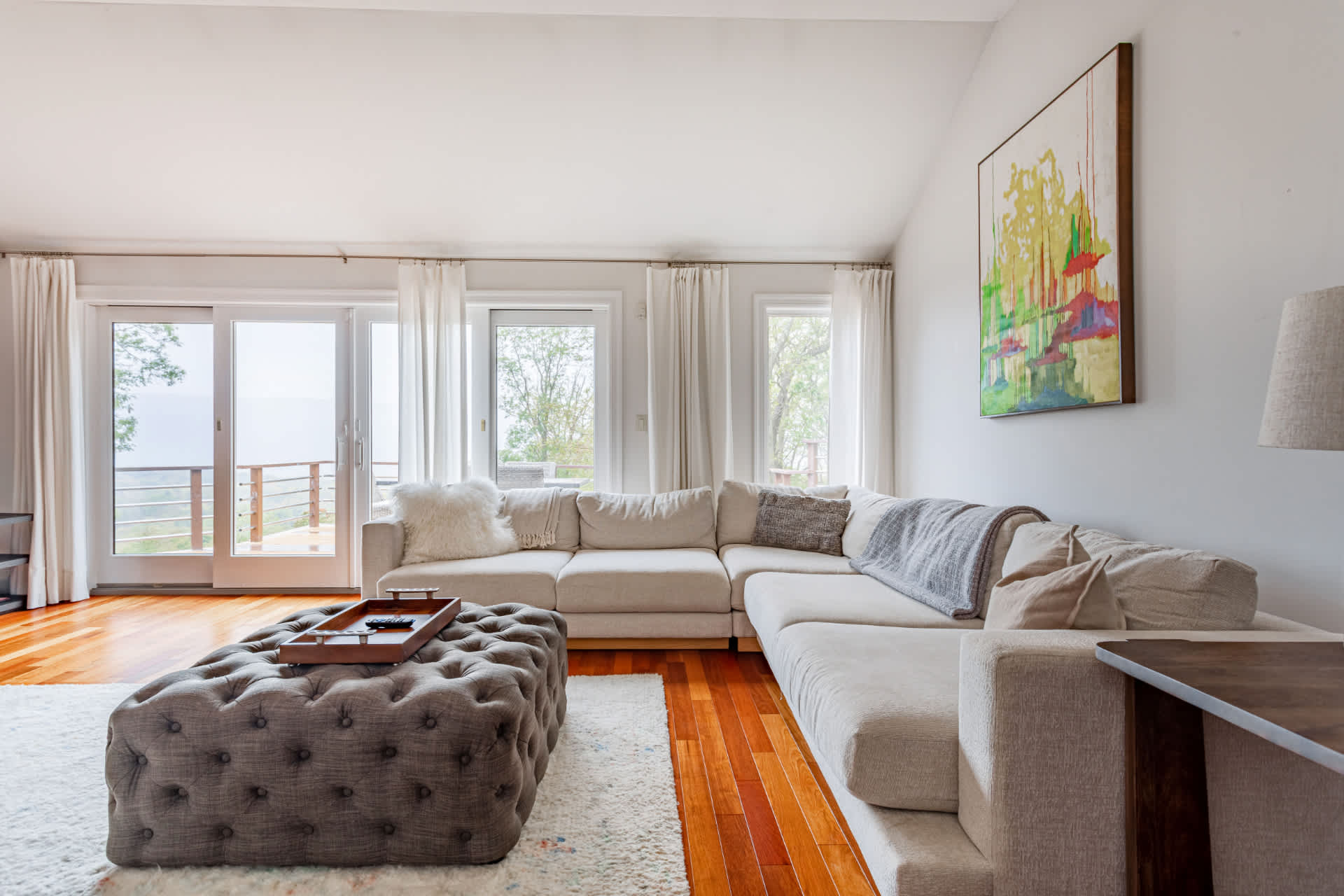 Bright living room with a large L-shaped sectional sofa and a tufted ottoman, facing expansive windows and sliding glass doors that offer a scenic outdoor view.