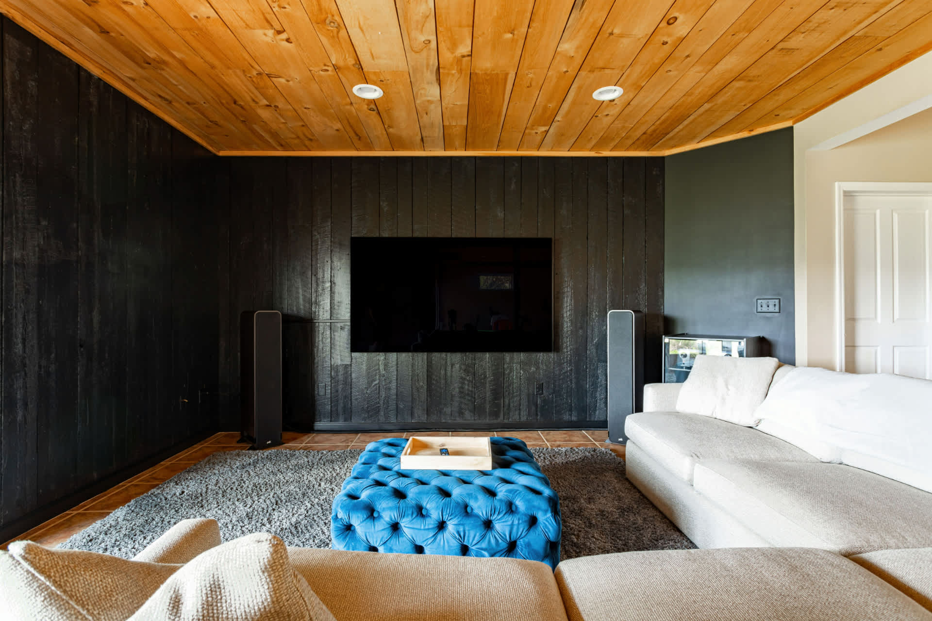 Cozy media room with dark wood paneled walls, a large wall-mounted TV, and a light-colored sectional sofa. A blue tufted ottoman serves as a coffee table, under a natural wood plank ceiling.