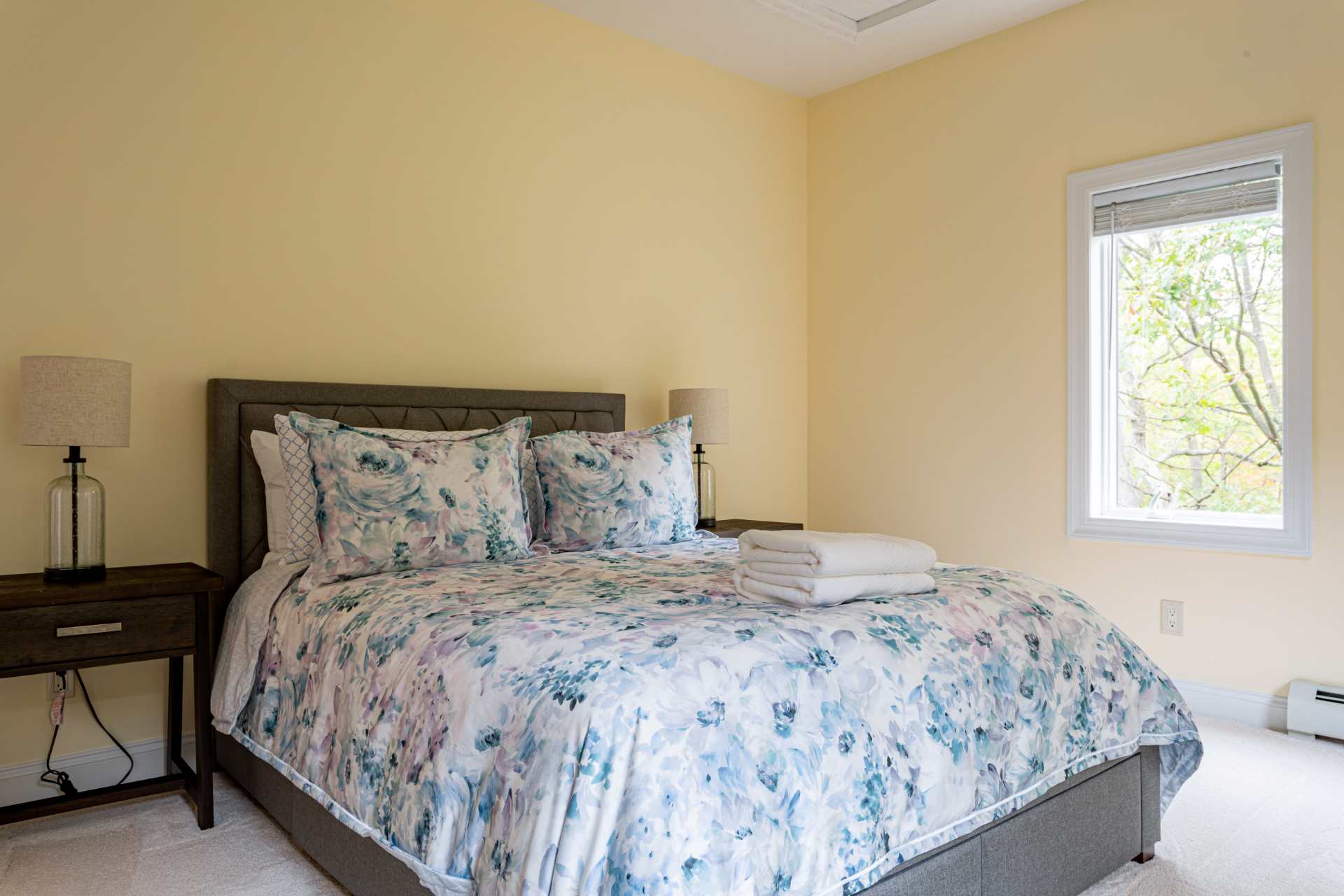 A cozy bedroom with light yellow walls, a queen-sized bed featuring a floral duvet and grey upholstered headboard, and a wooden nightstand with a lamp. A window provides natural light, and the floor is carpeted.