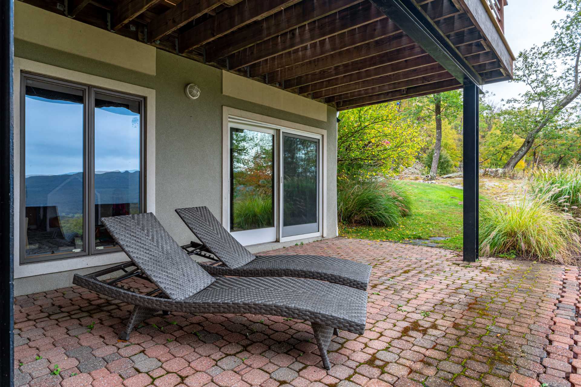 Ground-level outdoor patio with red paver stones, shaded by the deck above, featuring two dark woven lounge chairs and sliding glass doors leading indoors.