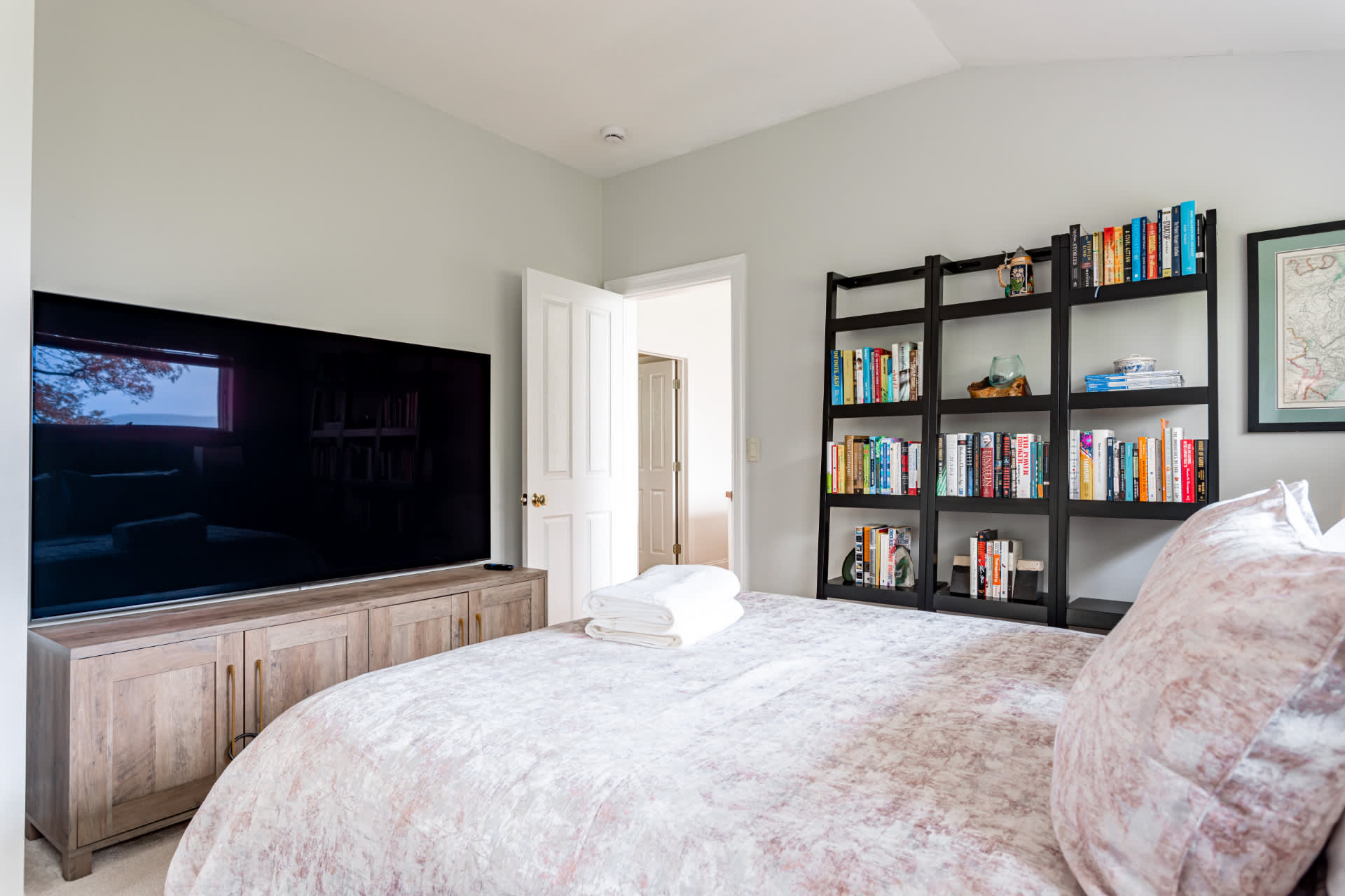 A bright bedroom featuring a large flat-screen TV on a rustic wooden console, a bed with a patterned duvet, and a tall black ladder-style bookshelf filled with books. A white door leads to an adjacent room.
