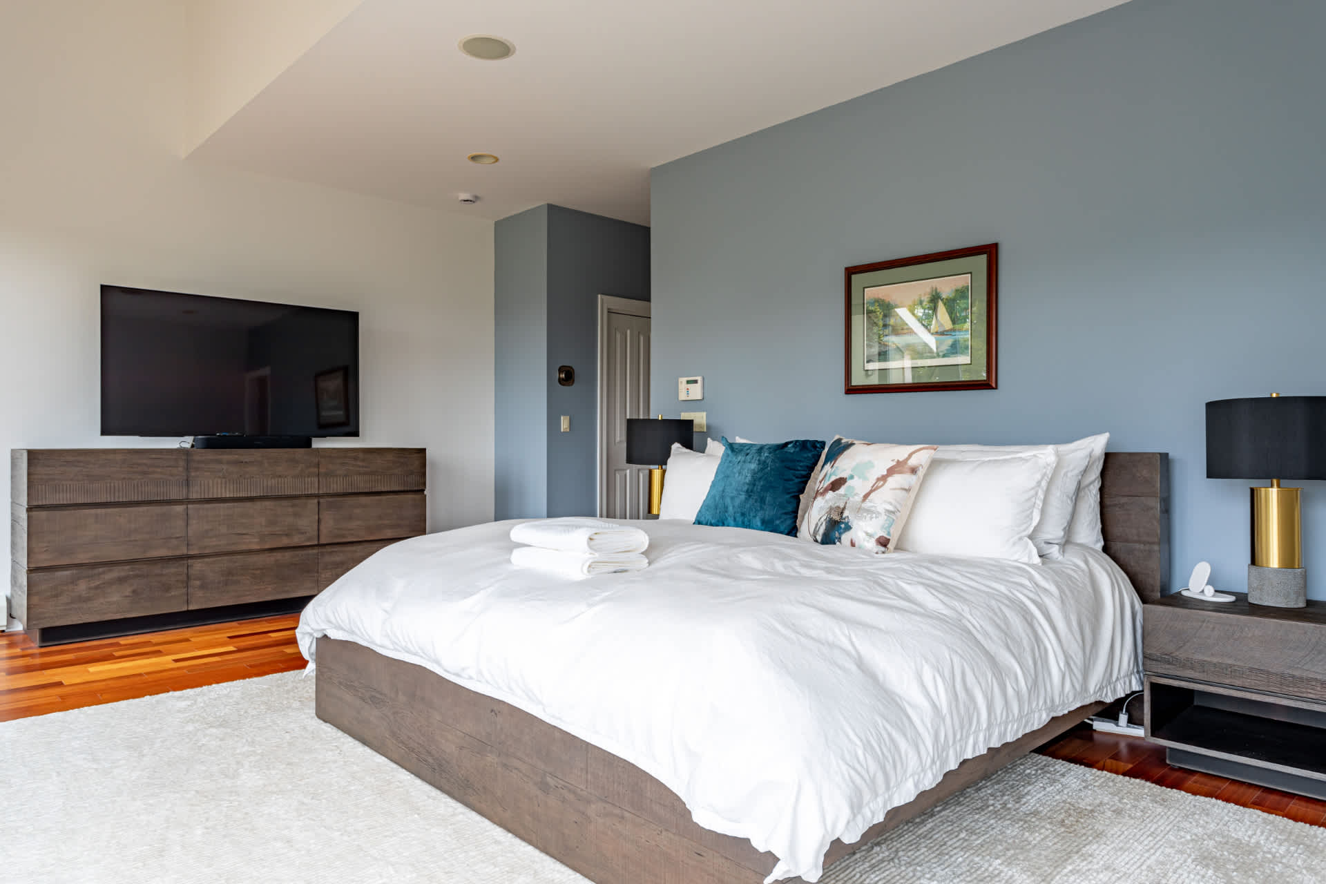 Spacious bedroom with a king-sized bed featuring a dark wood frame and white bedding, a large wall-mounted TV above a dresser, and light blue accent walls.