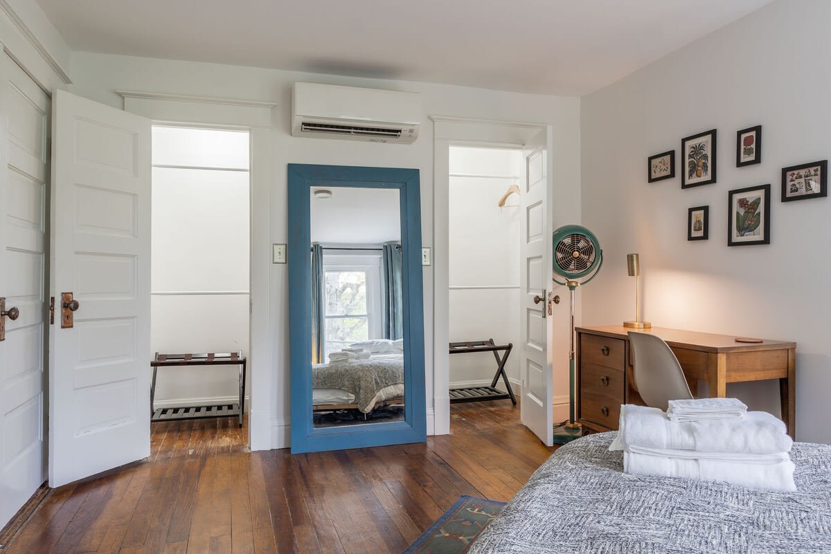A bright bedroom area with hardwood floors, featuring a wooden desk with a chair, a vintage-style fan, and a large blue-framed full-length mirror reflecting the bed and bay windows. Two open closet doors are visible.