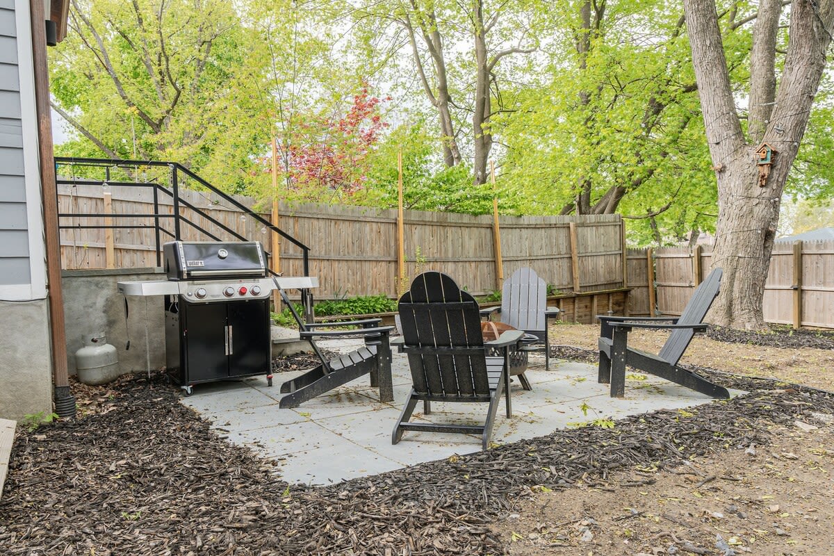 A spacious backyard patio features a black Weber BBQ grill next to a concrete slab with four black Adirondack chairs arranged around a small table, surrounded by wood chip landscaping and a wooden fence. Lush green trees provide a natural backdrop.