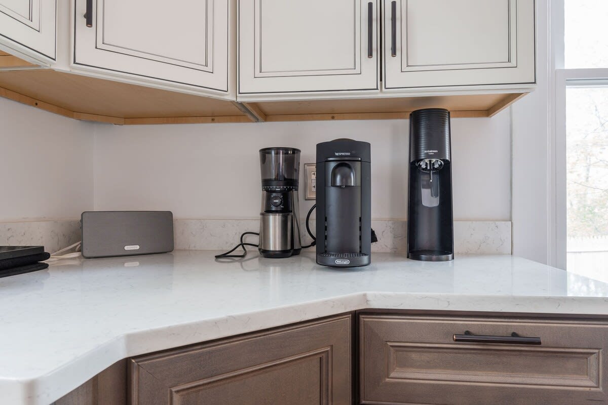 Detailed view of a kitchen counter with pristine white quartz countertops, showcasing a coffee grinder, a Nespresso machine, and a sparkling water dispenser. Two-tone cabinetry is visible above and below.