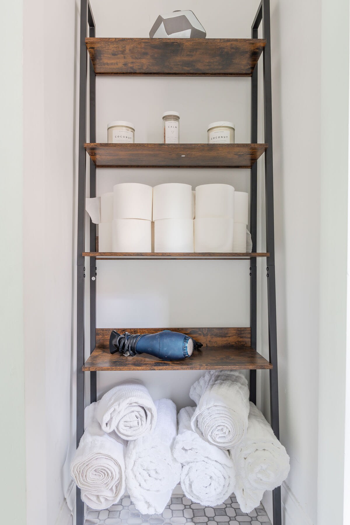 A tall, narrow shelving unit with dark metal frames and rustic wooden shelves, holding neatly stacked white toilet paper, rolled white towels, a blue hairdryer, and small candles. The bottom shelf has a white hexagonal tile floor.