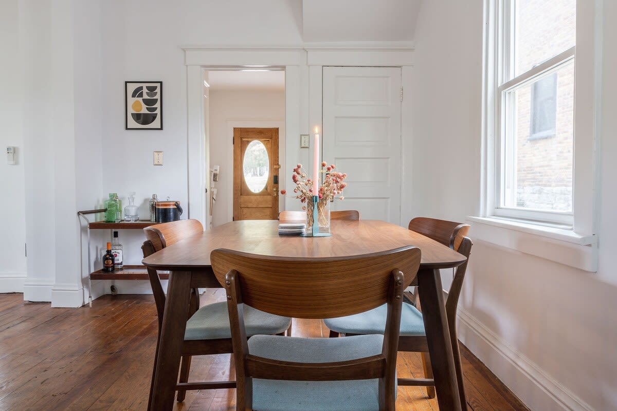 A cozy dining area with a wooden oval dining table and matching chairs with light blue cushions, set against white walls and hardwood floors, with a decorative bar cart in the corner.