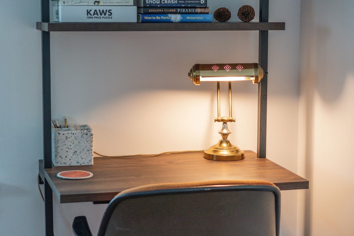 A dedicated workspace features a dark wooden desk with a vintage brass banker's lamp casting a warm glow, alongside a concrete pen holder and a coaster. Books are neatly stacked on the shelf above, suggesting a cozy reading or work nook.