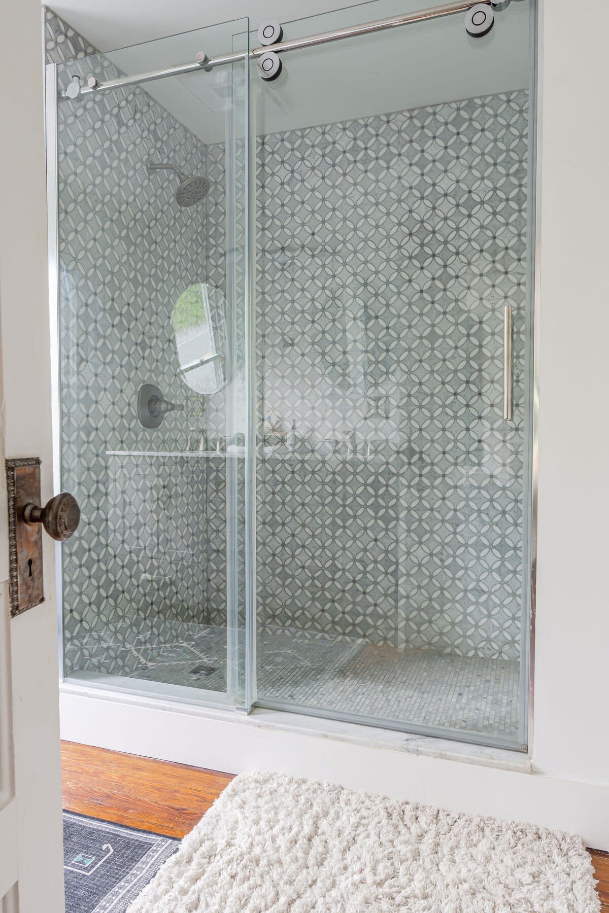 A modern shower enclosure with clear glass sliding doors, featuring light grey patterned tiles on the walls and small mosaic tiles on the floor, with a chrome showerhead and faucet.