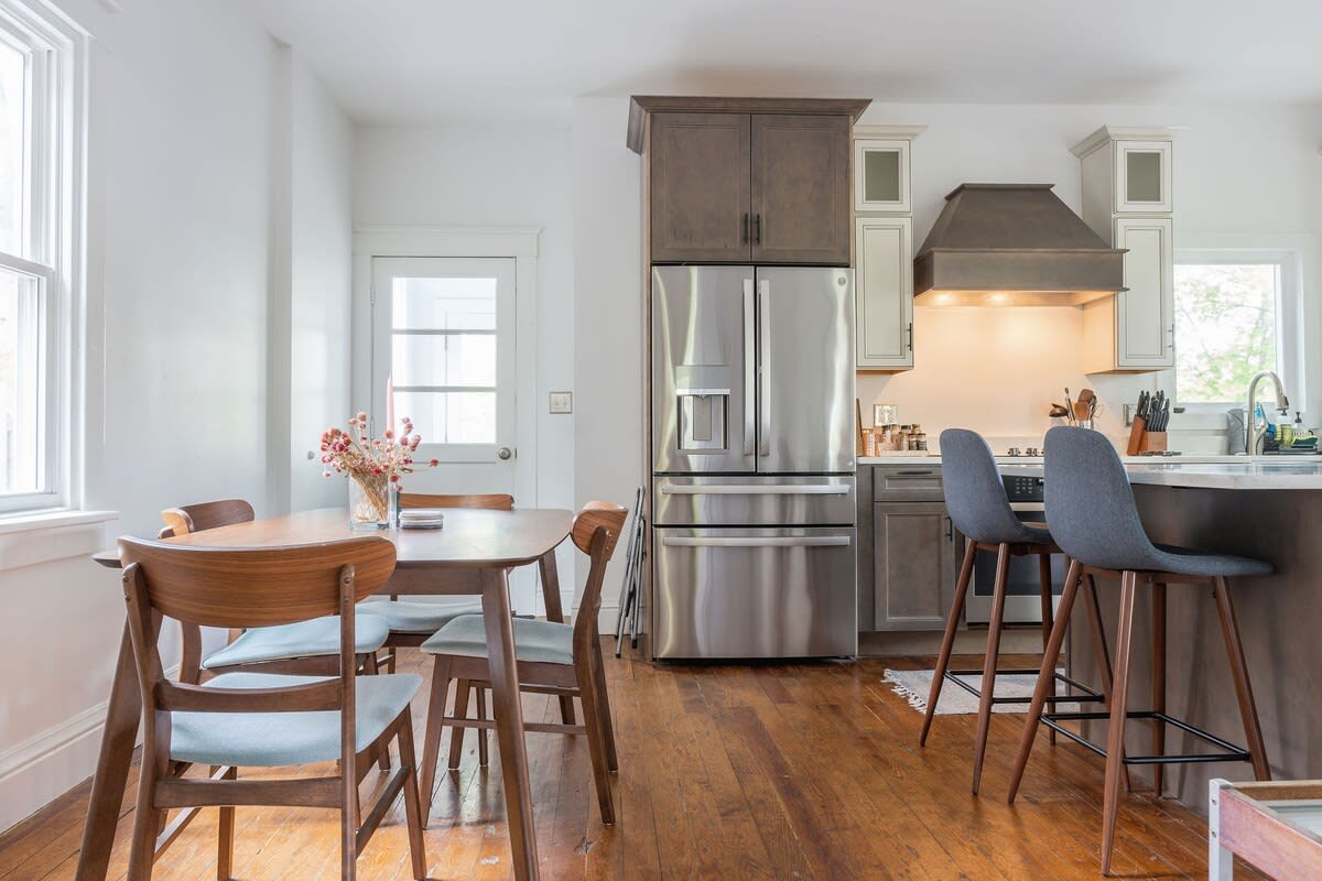 Open-concept dining area adjacent to the kitchen, furnished with a warm wooden dining table and four mid-century modern chairs. A stainless steel French door refrigerator and kitchen island are visible in the background.