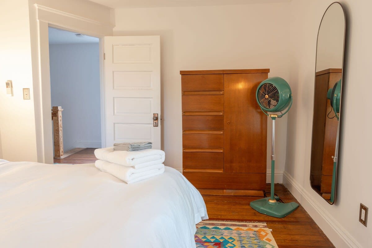 A view of a bedroom with hardwood floors, showing a wooden dresser with multiple drawers, a vintage green standing fan, and a full-length arched mirror reflecting the room.