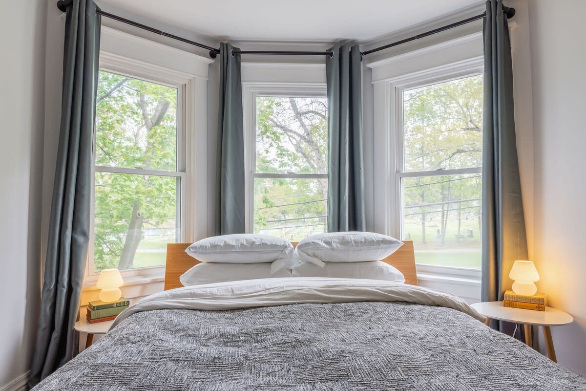 A cozy bedroom featuring a wooden headboard bed with white and grey bedding, flanked by two small side tables with lamps, set against a bay window with grey curtains and views of lush green trees.