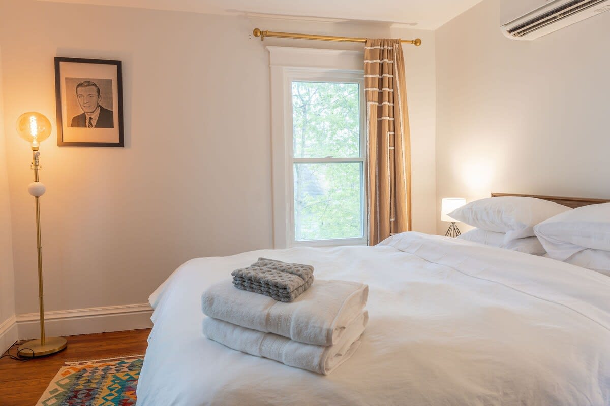 A bright bedroom featuring a bed with white bedding and folded towels, a single window with striped curtains, a tall floor lamp, and framed artwork on the wall.