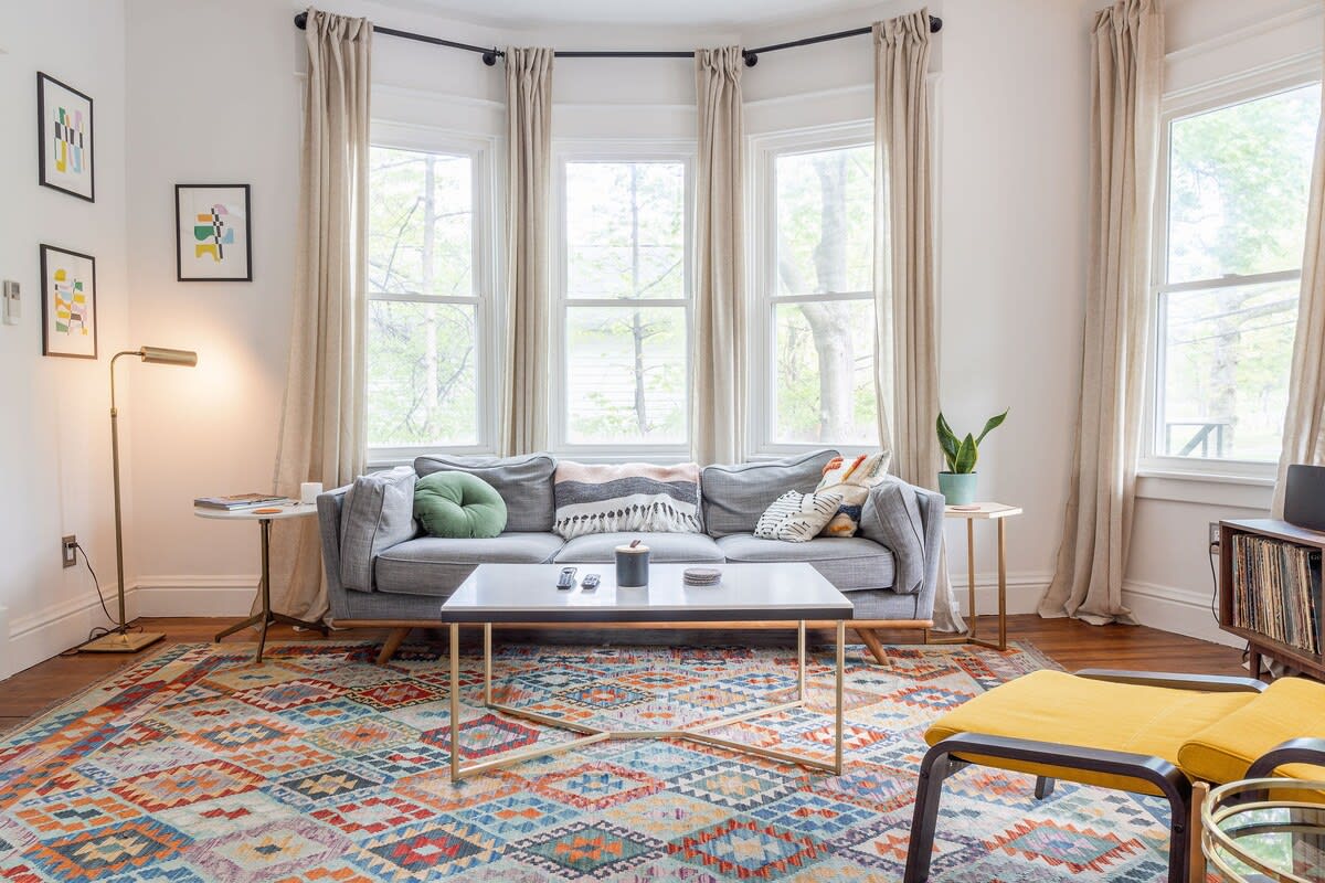 Bright living room featuring a gray sofa, a vibrant geometric rug, and a bay window with light-colored curtains. A modern coffee table with a gold base and a brass floor lamp complete the space.