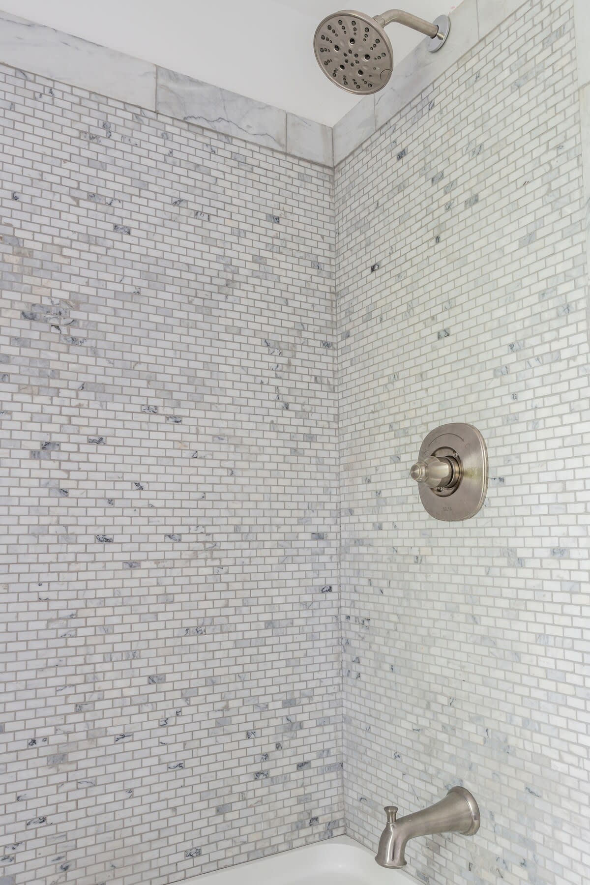 A bright shower/tub area with white subway-style rectangular mosaic tiles on the walls, featuring a chrome showerhead and tub faucet, and a white bathtub.