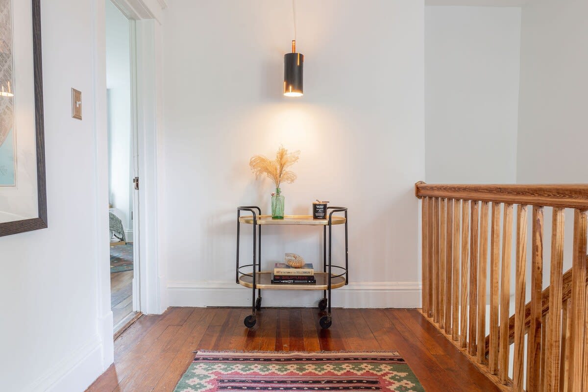A bright hallway with polished hardwood floors features a modern black and gold bar cart adorned with a vase of dried pampas grass and books, illuminated by a sleek black and gold wall sconce. A colorful patterned rug lies on the floor, leading towards an open doorway.