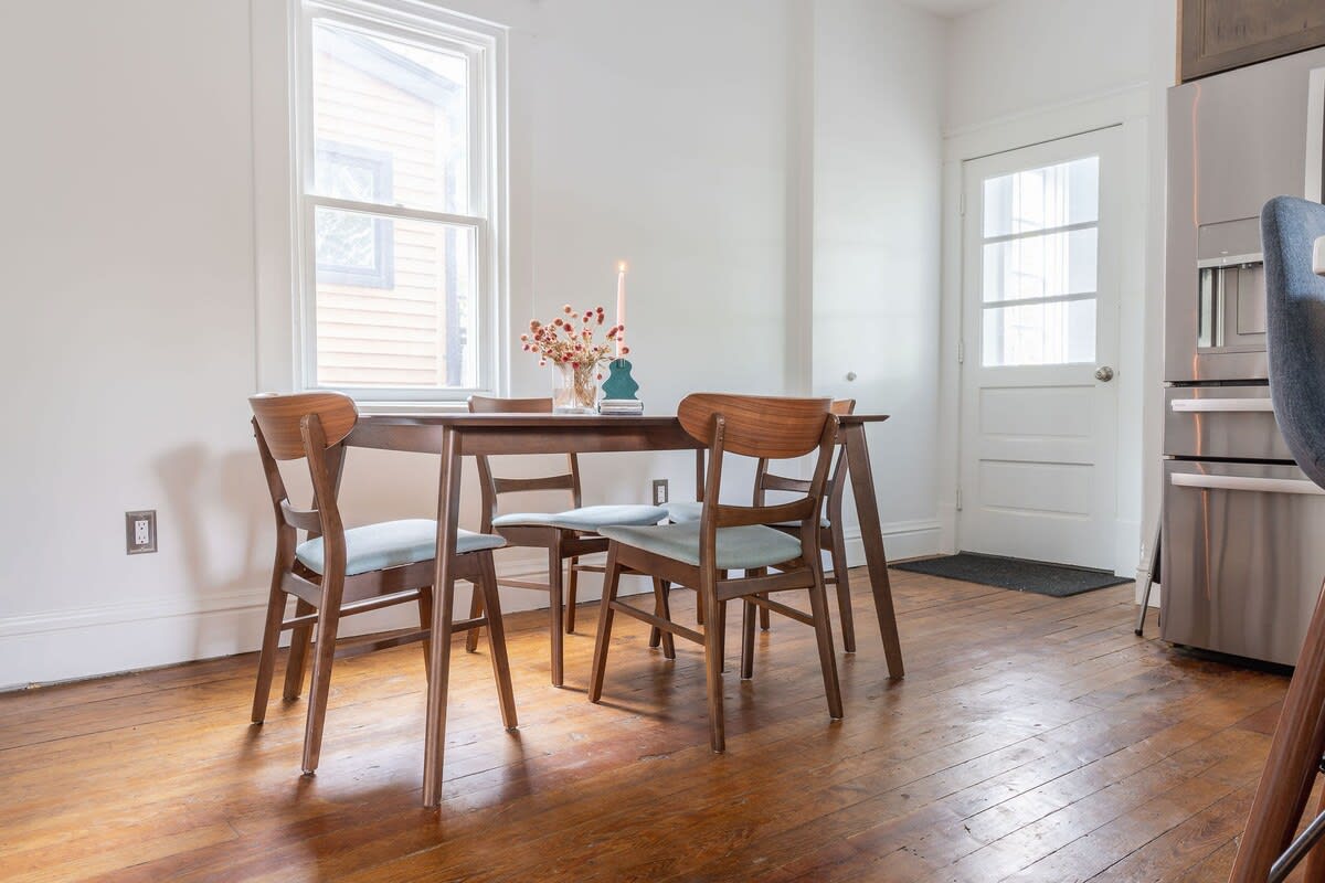 Bright dining area featuring a wooden table and four chairs, positioned by a large window. A white door with a glass panel leads outside, and a vase with dried flowers serves as a centerpiece.