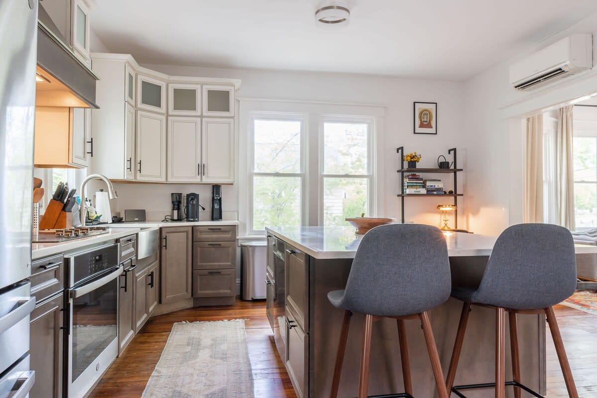 Modern kitchen featuring two-tone cabinetry with white upper and gray lower cabinets, stainless steel appliances, and a large island with two gray upholstered bar stools. A window offers natural light, and an AC unit is mounted above.