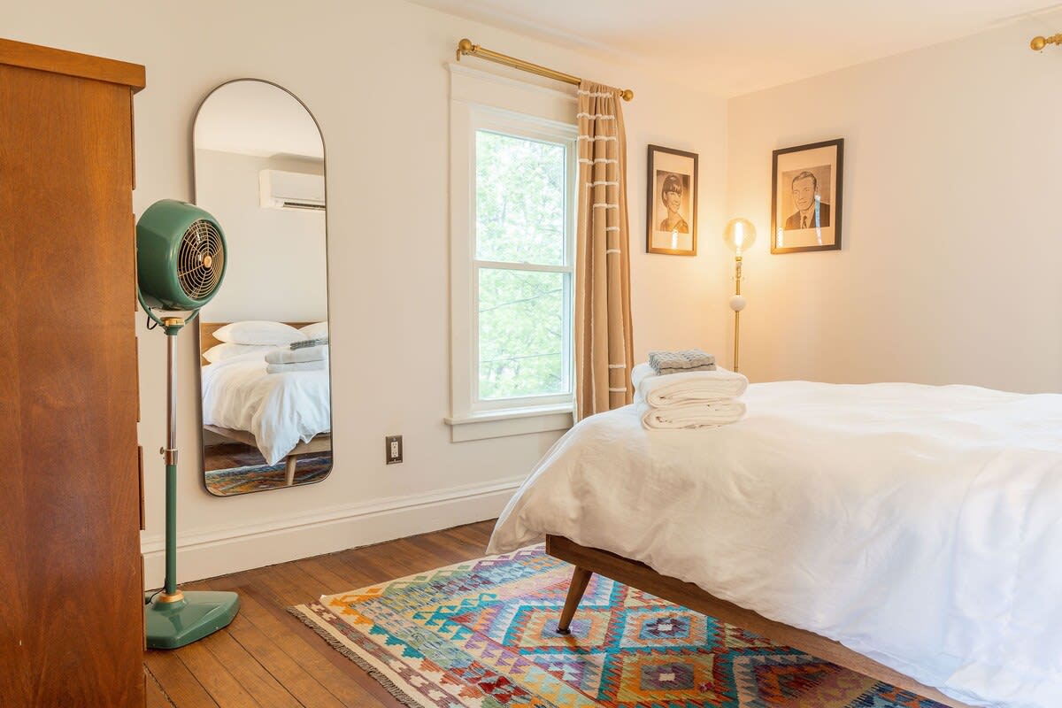 A cozy bedroom with hardwood floors and a colorful geometric rug, featuring a bed with white bedding, a tall arched mirror, a vintage green standing fan, and a window with striped curtains.