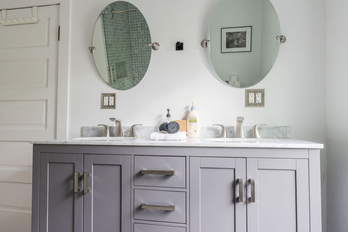 A contemporary bathroom vanity with a grey cabinet, white marble countertop, and two oval sinks with brushed nickel faucets, flanked by two oval mirrors and wall-mounted light fixtures.