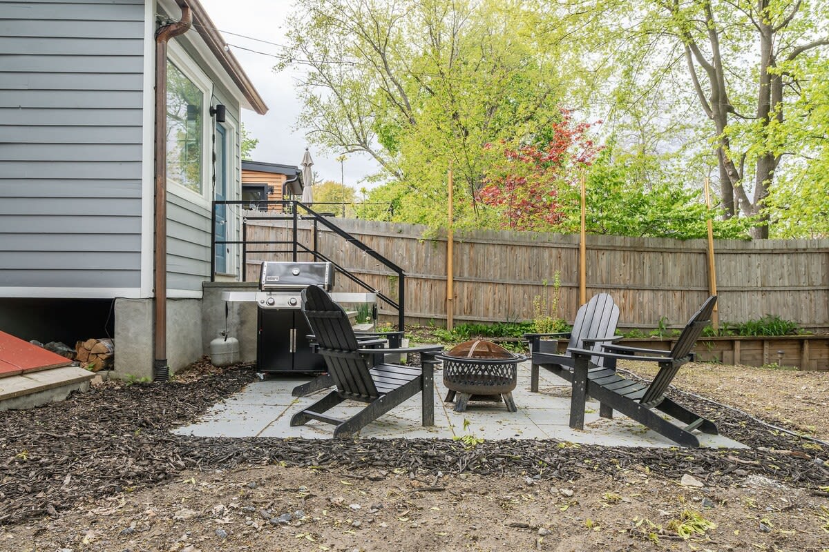 A backyard patio area with a fire pit surrounded by two black Adirondack chairs, a BBQ grill, and a wooden fence, offering a space for outdoor relaxation.