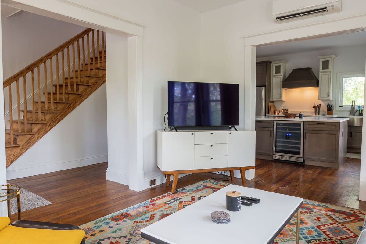 A view from the living room towards the kitchen and staircase, featuring a large flat-screen TV on a white media console, and a colorful rug on warm hardwood floors.