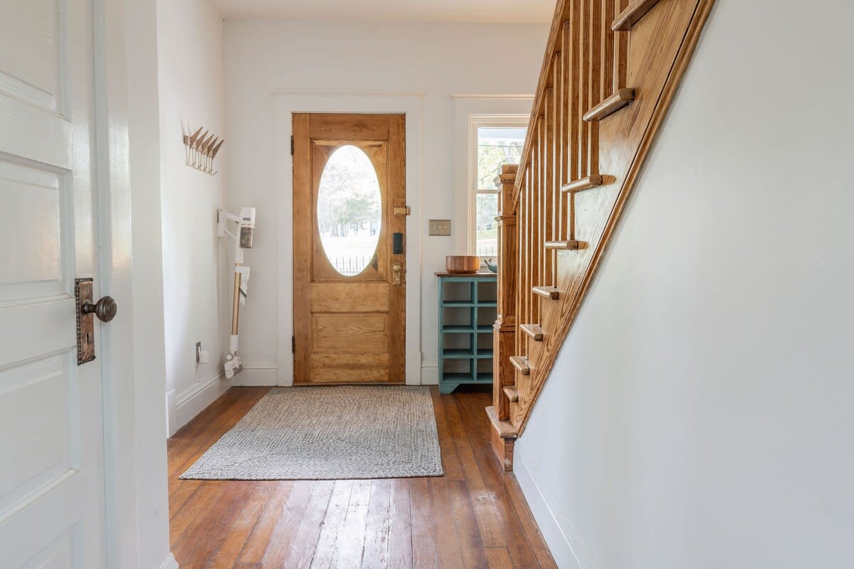 Inviting entryway with a natural wood front door featuring an oval glass insert, and a light gray rug on hardwood floors. A wooden staircase with a banister leads upstairs, and a coat rack is mounted on the wall.