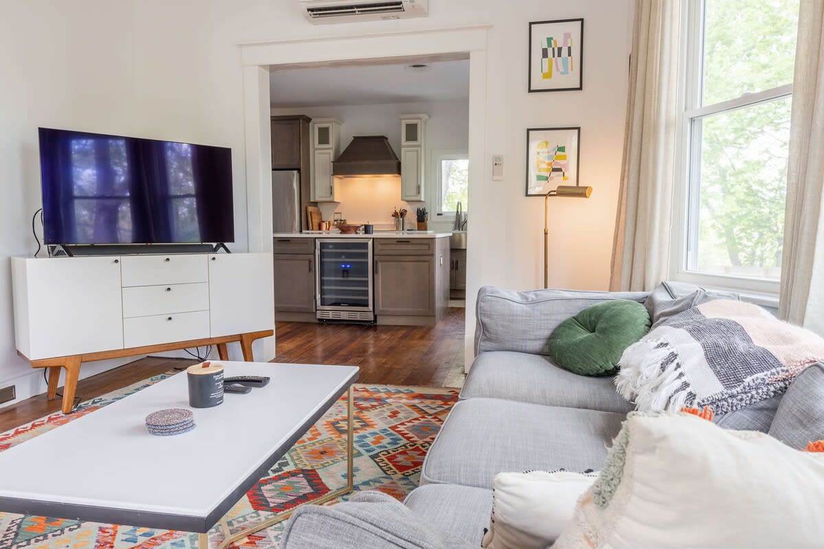 View of the living room with a large flat-screen TV on a white media console, facing a comfortable gray sectional sofa. An open archway provides a glimpse into the adjacent kitchen with a wine fridge.