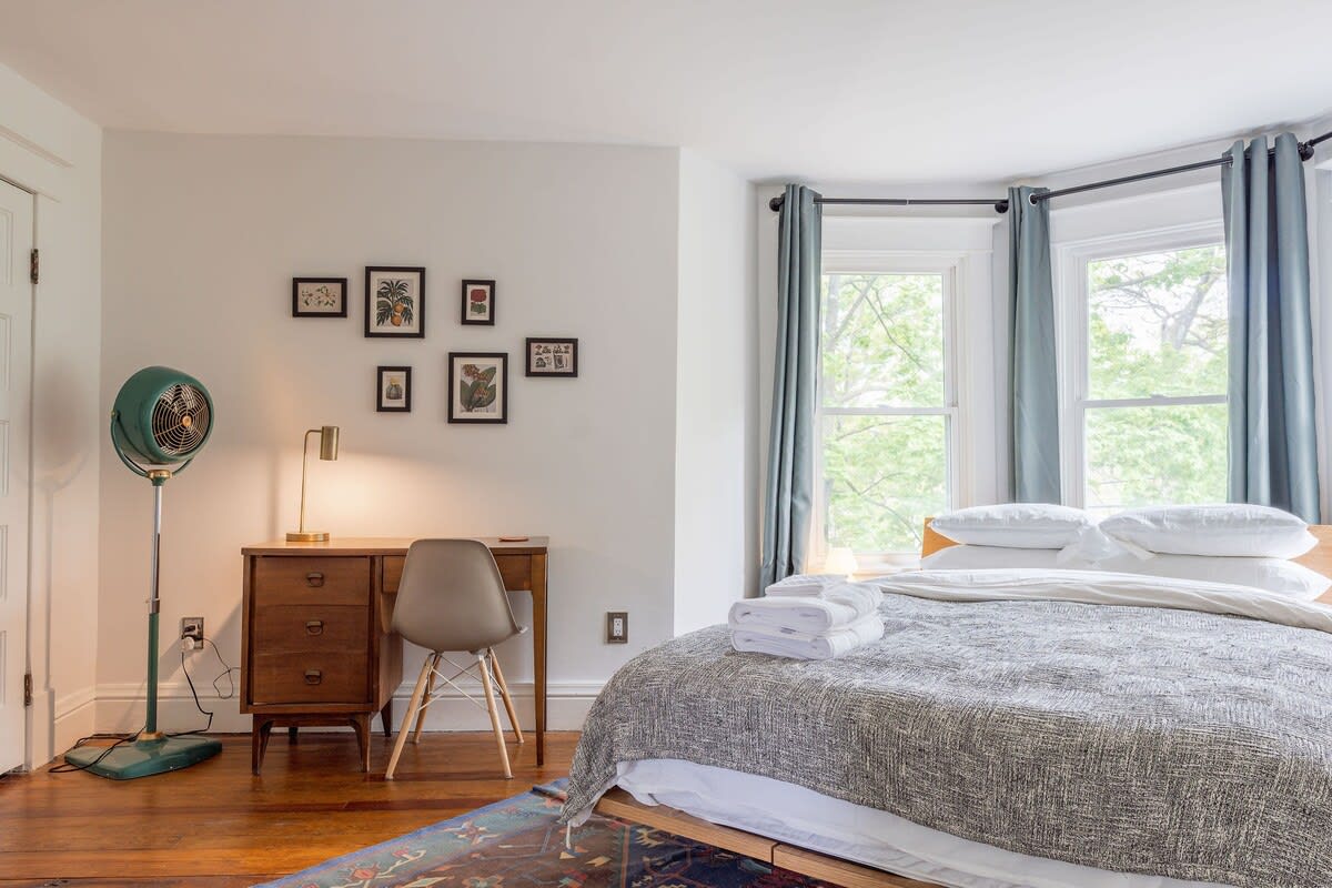 Cozy bedroom featuring a queen-sized bed with a gray textured blanket, light blue curtains on bay windows, and a vintage-style green floor fan. A wooden desk with a task lamp and framed artwork adorns the wall.