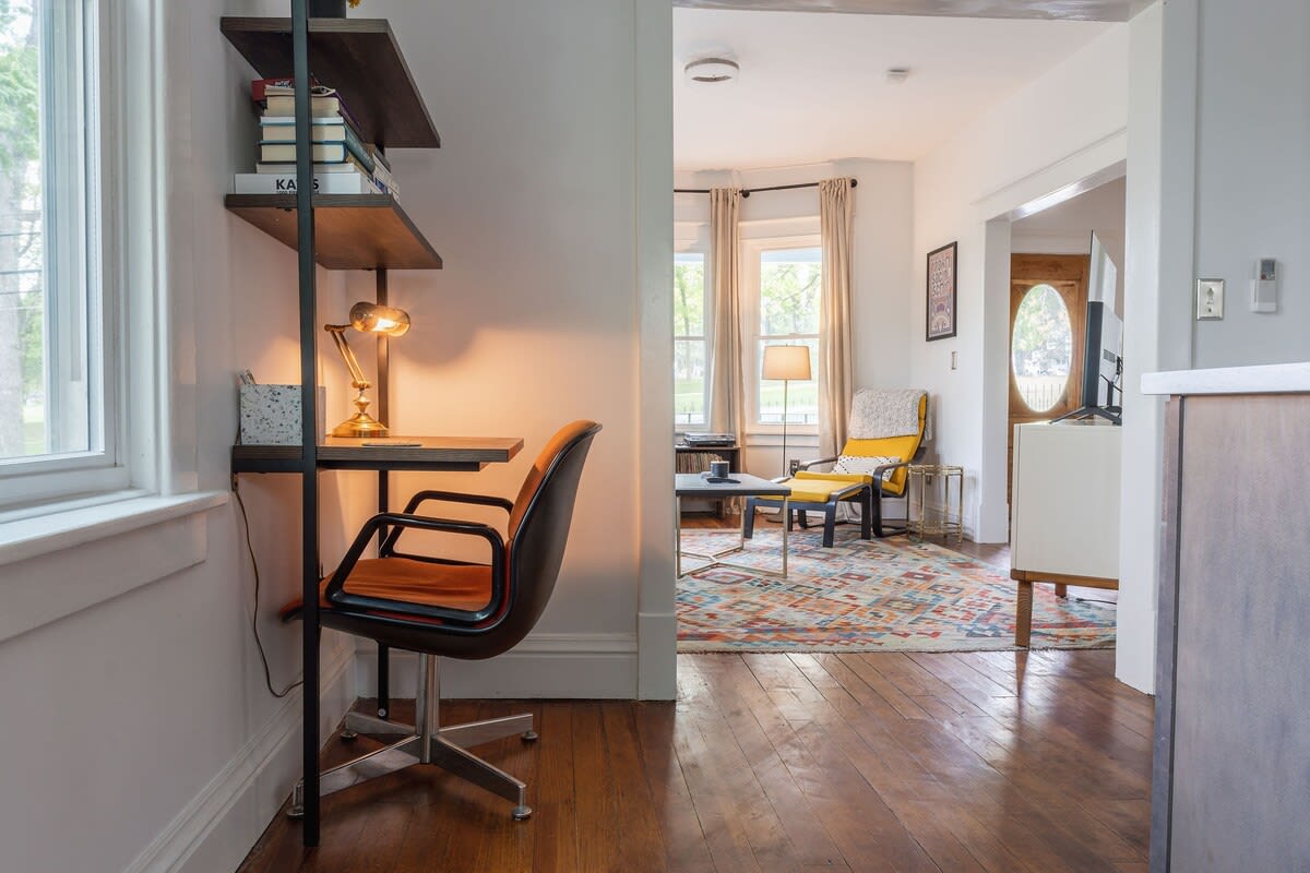 A dedicated workspace with a modern desk and chair, integrated shelving, and a task lamp, offering a view into the living room and natural light from a nearby window.