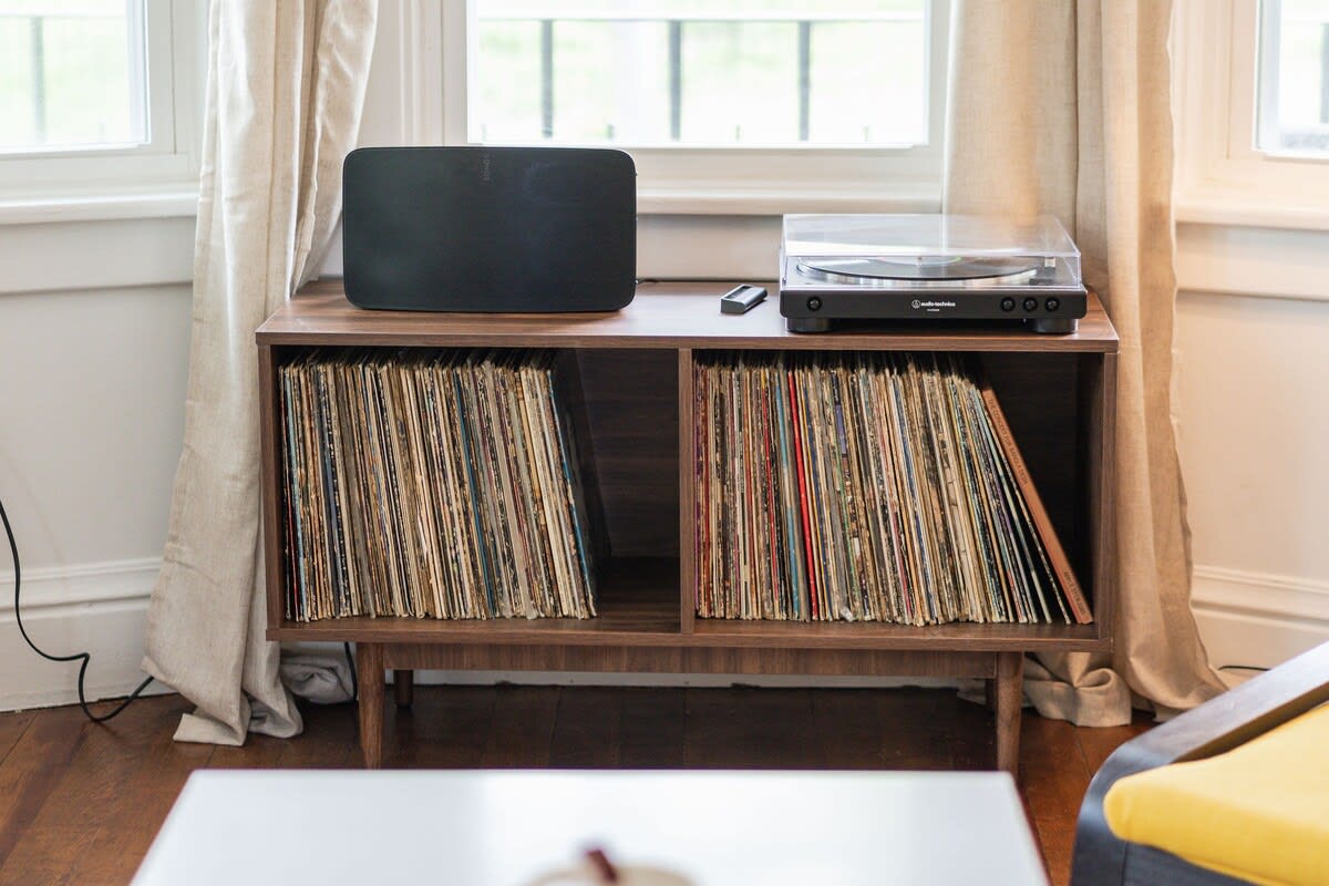 Close-up of a mid-century modern record console filled with vinyl records, topped with a turntable and a black Sonos speaker. It is positioned by a window with light drapes.