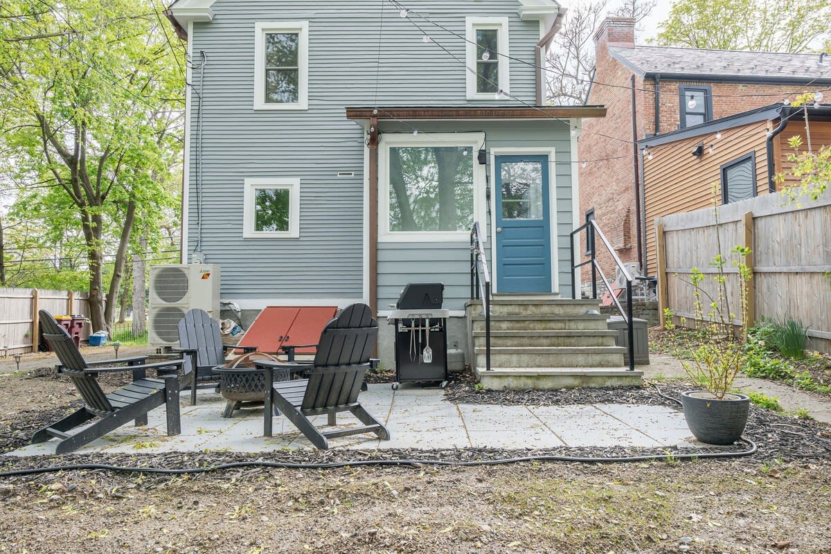 The back of the light blue house features a small patio area with black Adirondack chairs and a BBQ grill, accessible via concrete steps leading to a blue back door. An outdoor AC unit is visible, and a wooden fence encloses the yard.