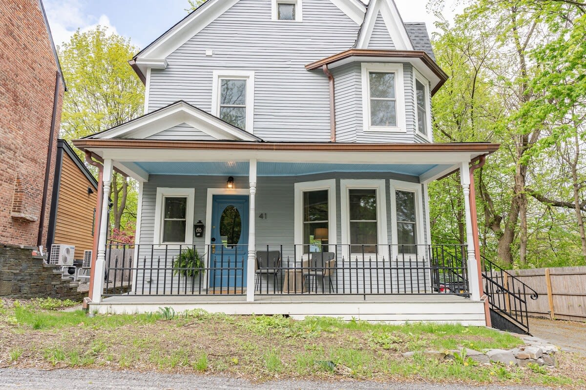 A detailed view of the light blue Victorian house's front facade, showcasing its inviting porch with black railings, a vibrant blue front door, and two dark chairs. The porch ceiling is painted a light blue, complementing the house siding.