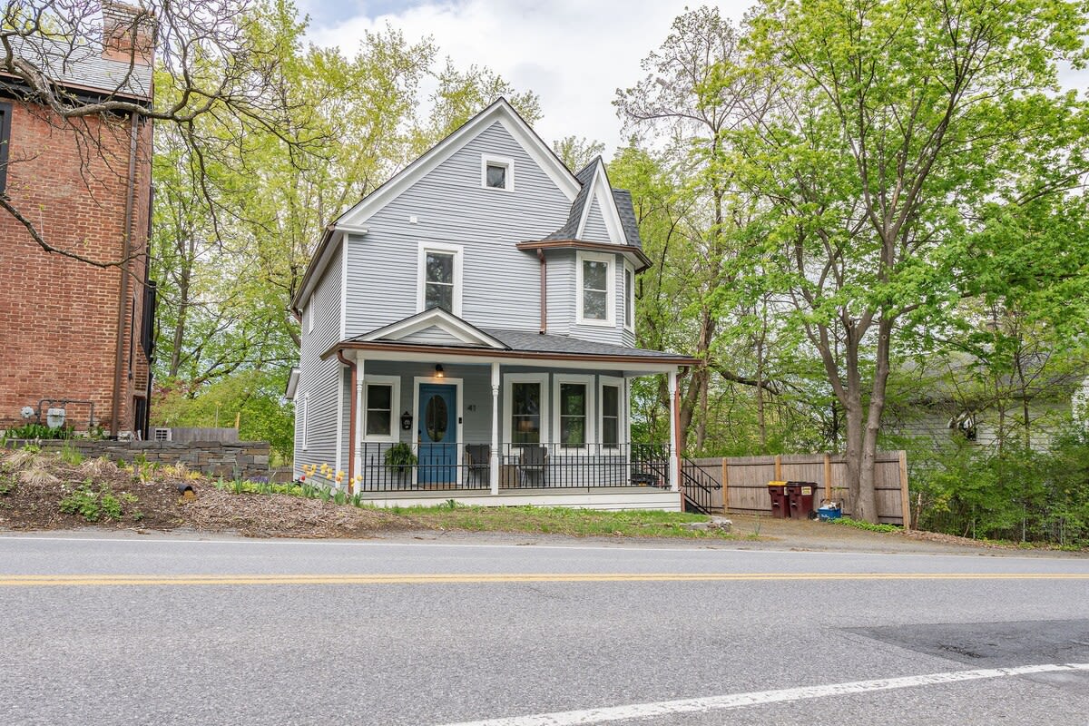 A charming light blue Victorian-style house with white trim and a dark roof stands prominently by a road, featuring a welcoming front porch with a bright blue door and multiple windows. Mature green trees frame the property, enhancing its curb appeal.